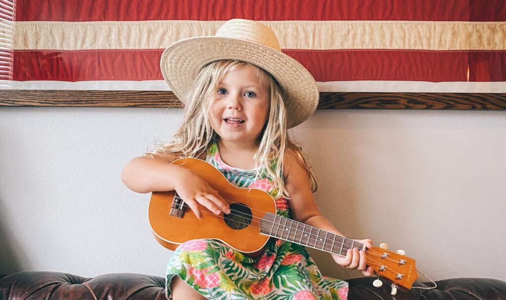 Little girl sitting in front American Flag wearing dress and hat, and holding guitar